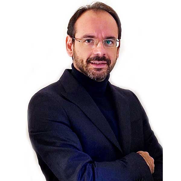 Un uomo in un blazer scuro e dolcevita posa con le braccia incrociate su uno sfondo bianco.