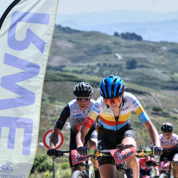 Two cyclists compete in a mountain biking event, with one in a blue helmet and colourful jersey, and a large flag in the background.