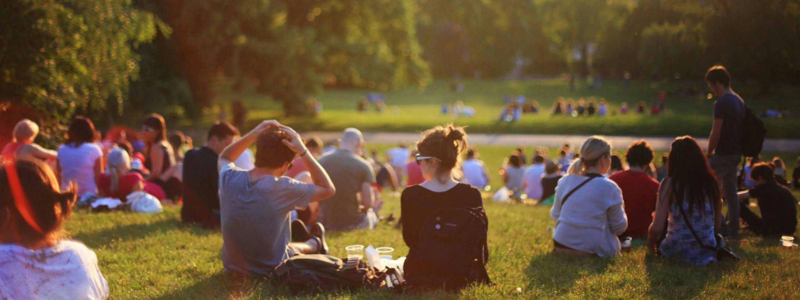 Una folla di persone sedute sull'erba in un parco, godendosi una giornata di sole con alcune tazze nei pressi.