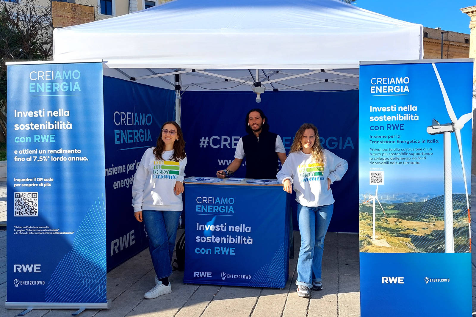 Uno stand promozionale con due donne in felpe e un uomo dietro a un tavolo, con messaggi sulla sostenibilità e codici QR.