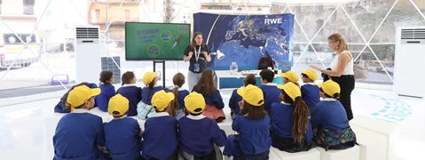 Gruppo di bambini con cappelli gialli che osservano attentamente una presentazione sul cambiamento climatico all'interno di una cupola geodetica.