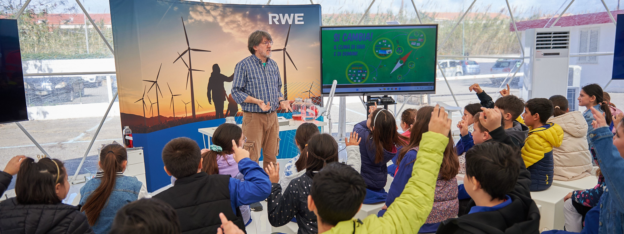 Un insegnante presenta ai bambini in una tenda con turbine eoliche e schermi tecnologici, molti studenti alzano le mani.