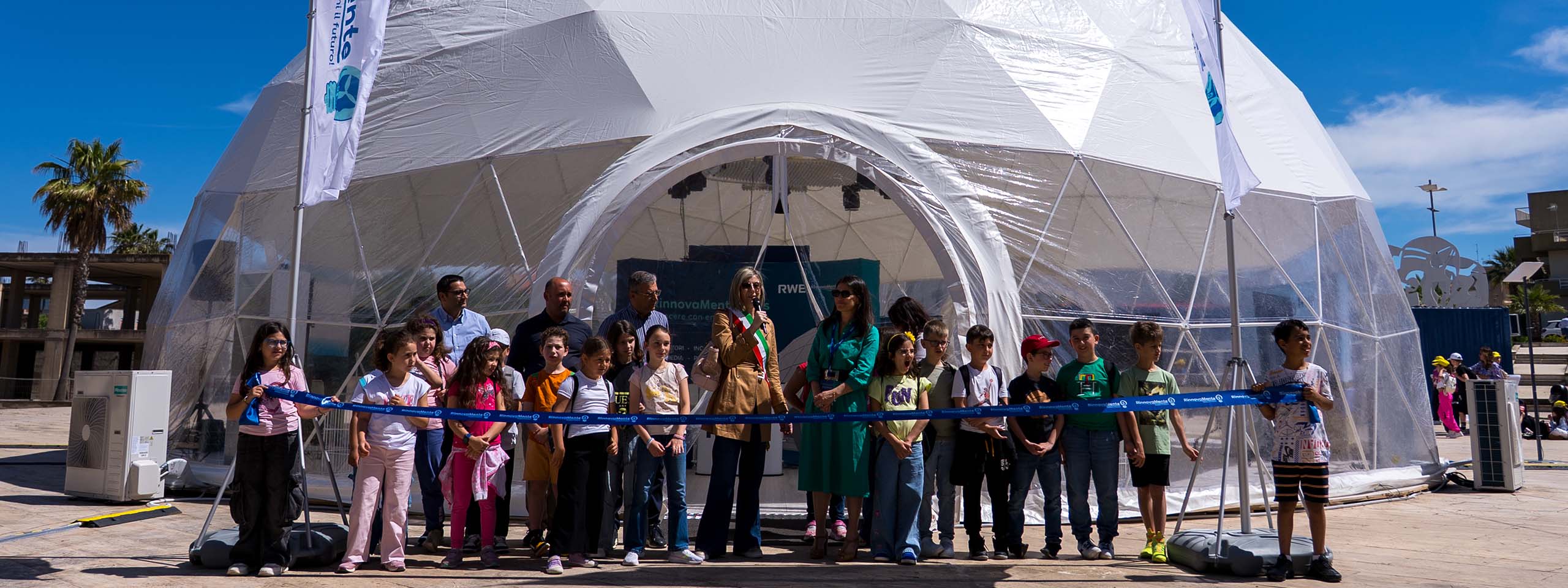 Un gruppo di bambini e adulti in fila tiene un lungo nastro blu davanti all'ingresso di una cupola geodetica bianca in tessuto, con cielo azzurro e palme sullo sfondo.