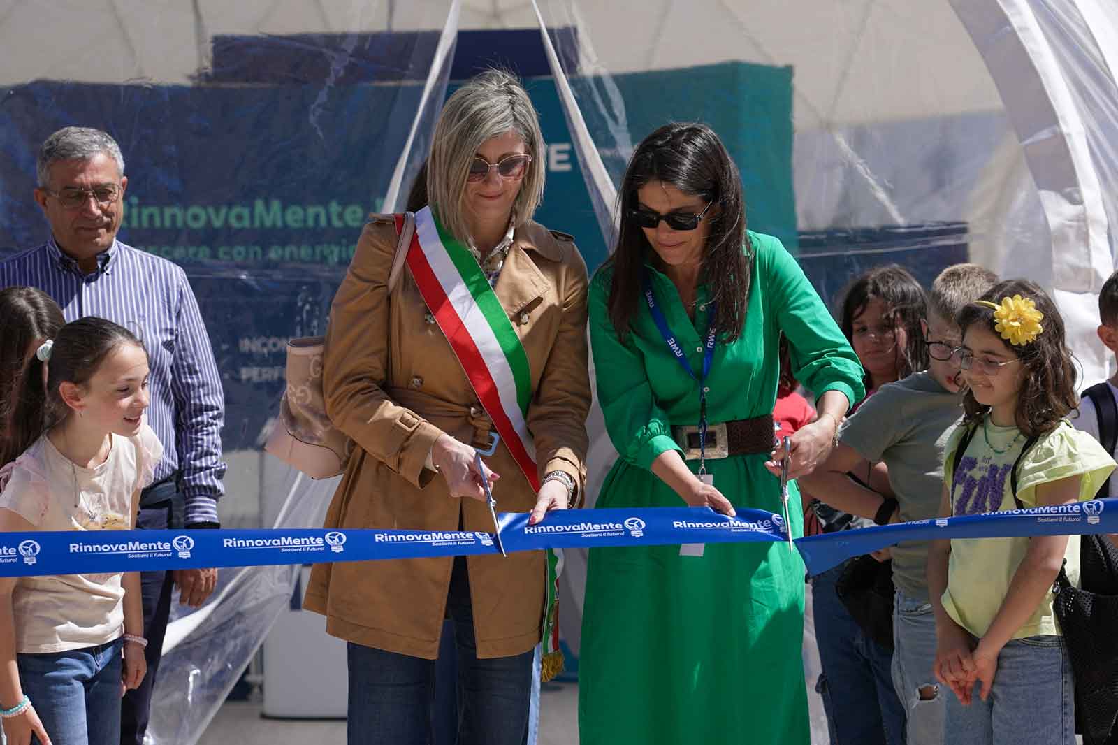 Due donne tagliano un nastro blu a un evento, circondate da bambini e adulti in un'atmosfera festosa.