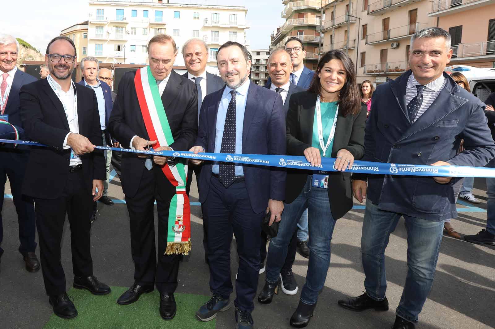 Un gruppo di funzionari, incluso un uomo con una fascia cerimoniale, taglia un nastro blu durante un evento pubblico.