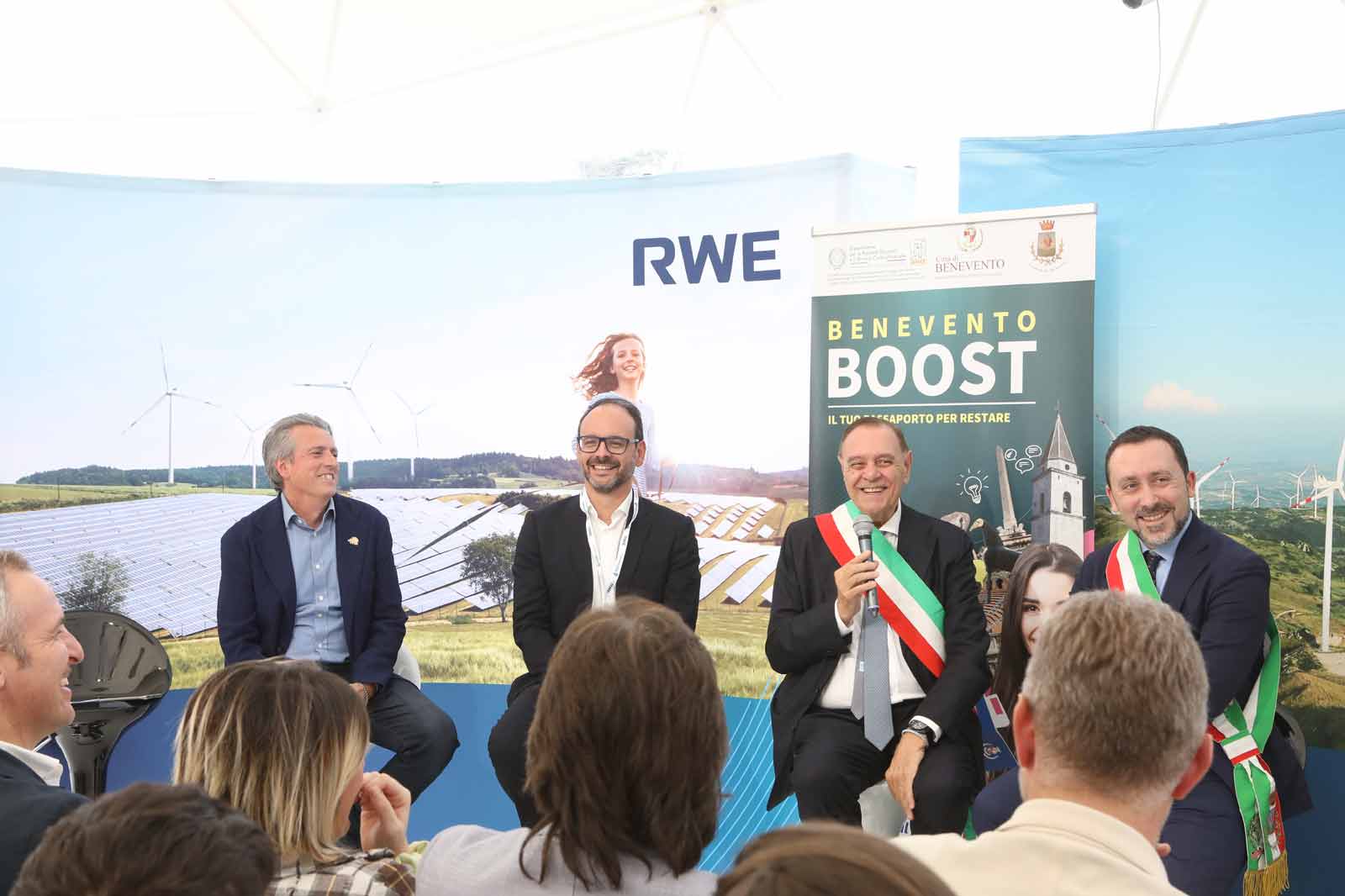 Un gruppo di persone sedute a un evento, discutendo argomenti legati all'energia rinnovabile con pannelli solari e turbine eoliche sullo sfondo.