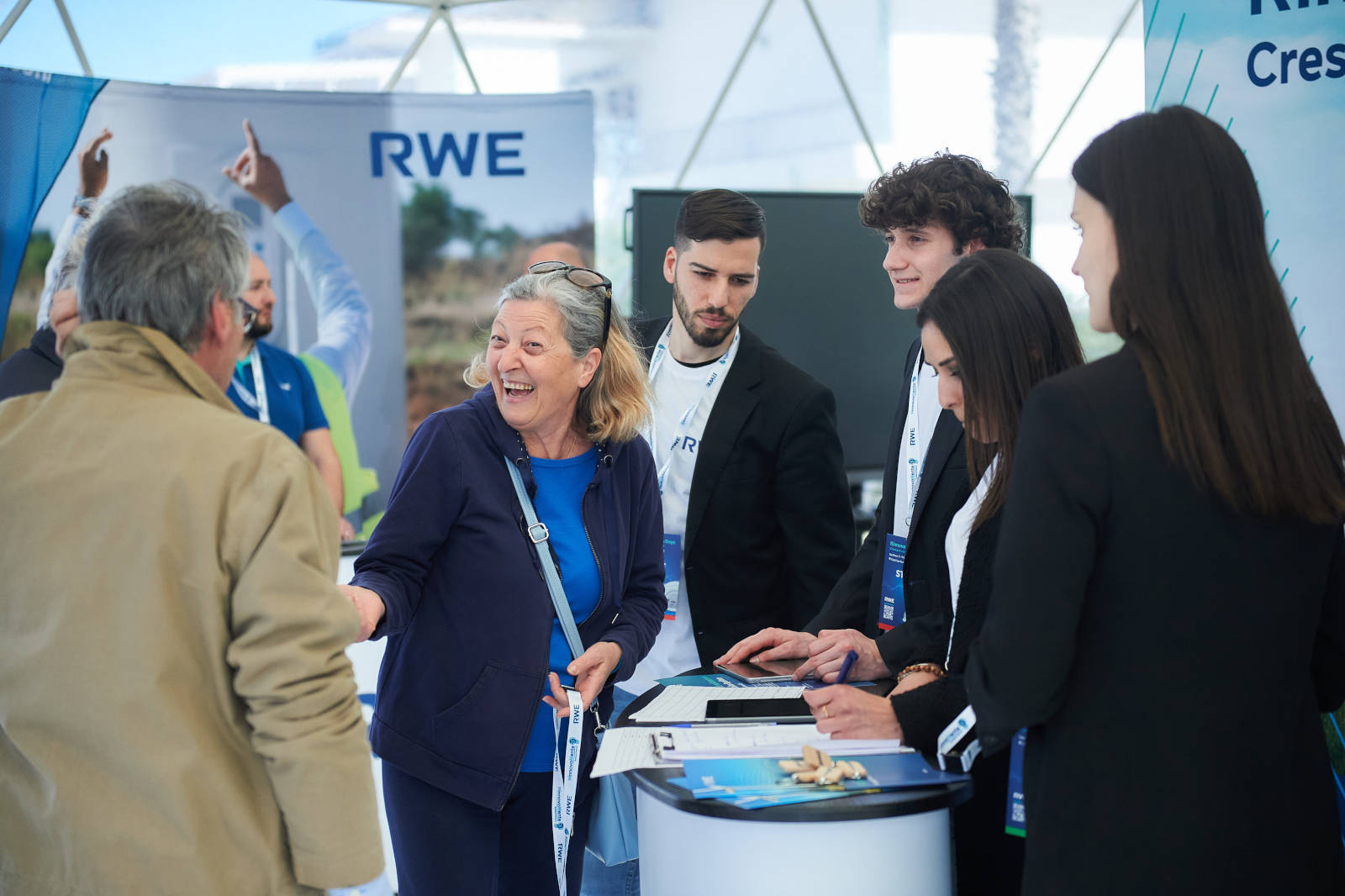 Un gruppo di persone che discutono presso uno stand RWE. Sono visibili espositori e materiali promozionali.