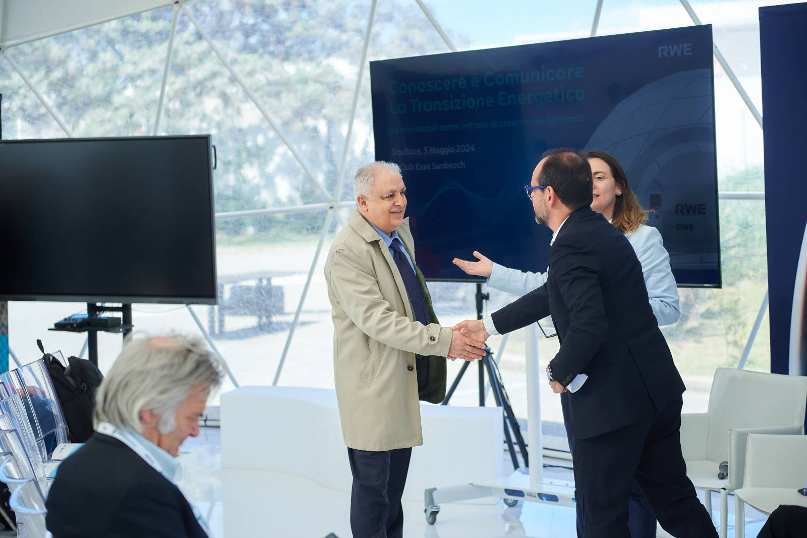 Due uomini si stringono la mano a una conferenza, discutendo della transizione energetica, con una presentazione sullo sfondo.