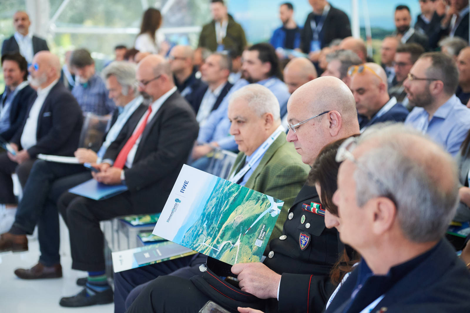 Un pubblico di persone sedute in una sala conferenze, alcuni con opuscoli. Il contesto è professionale e formale.