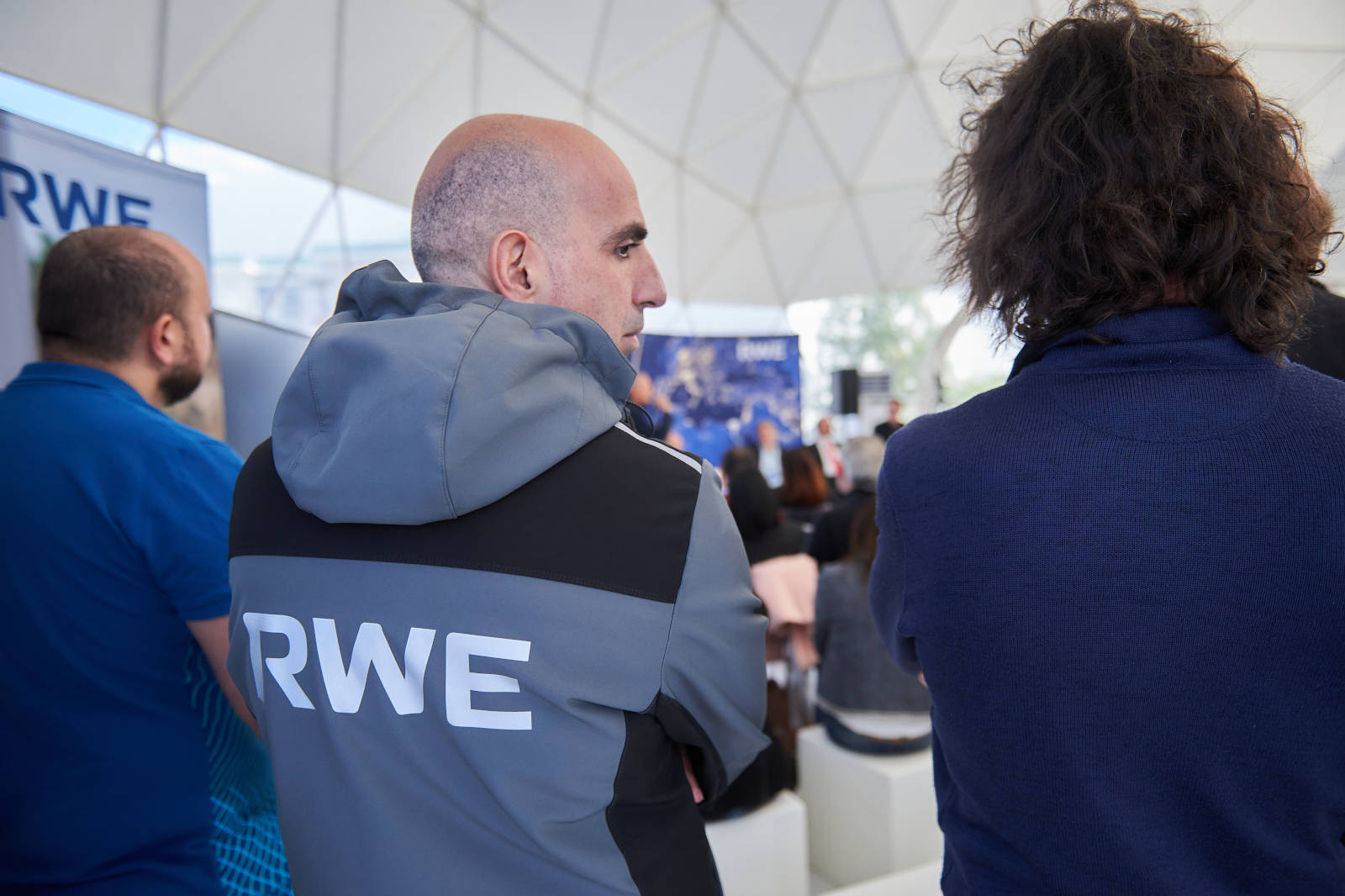 Un gruppo di persone che osservano una presentazione sotto una struttura a cupola geodetica, con un focus su una persona in giacca con 'RWE' sulla schiena.