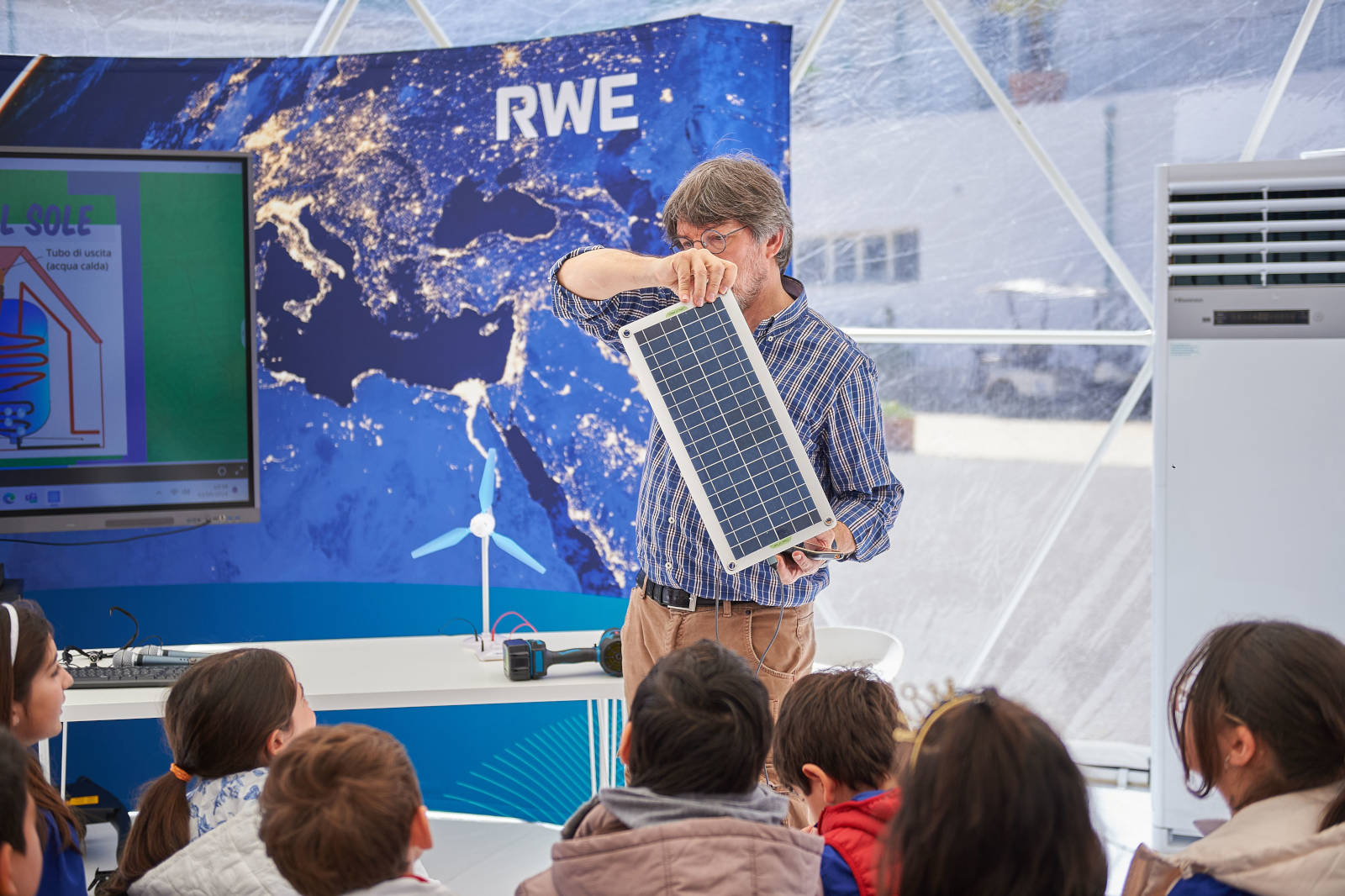 Un uomo dimostra un piccolo pannello solare ai bambini, con un modello di turbina eolica e materiali educativi sullo sfondo.