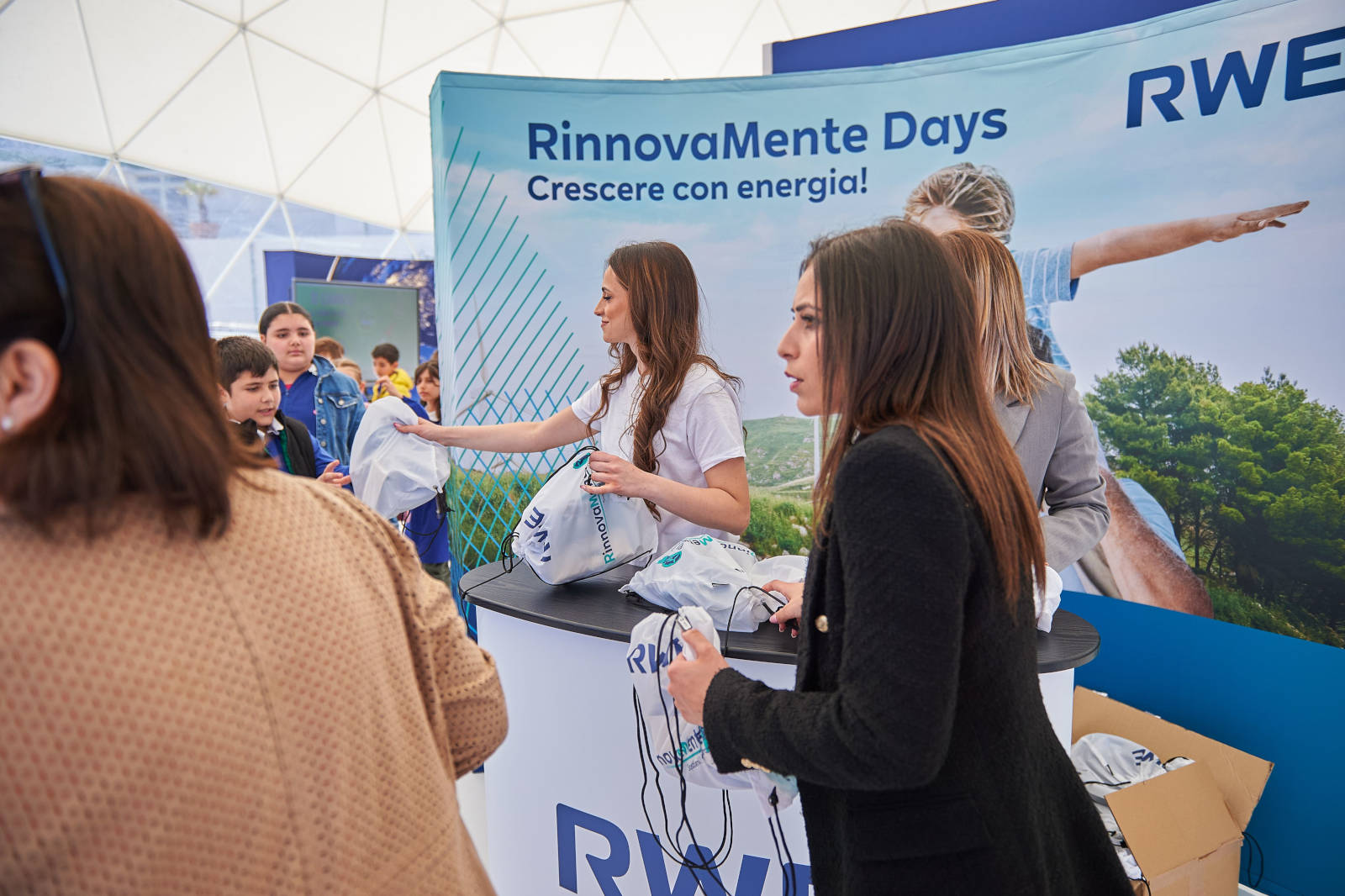 Un evento con persone che ricevono borse promozionali da uno stand RWE. Banner con 'RinnovaMente Days'.
