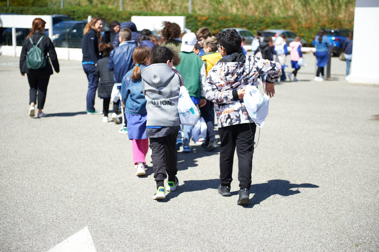 Un gruppo di bambini che cammina su una superficie pavimentata, alcuni tengono borse, vestiti in abbigliamento casual, sotto la luce solare.