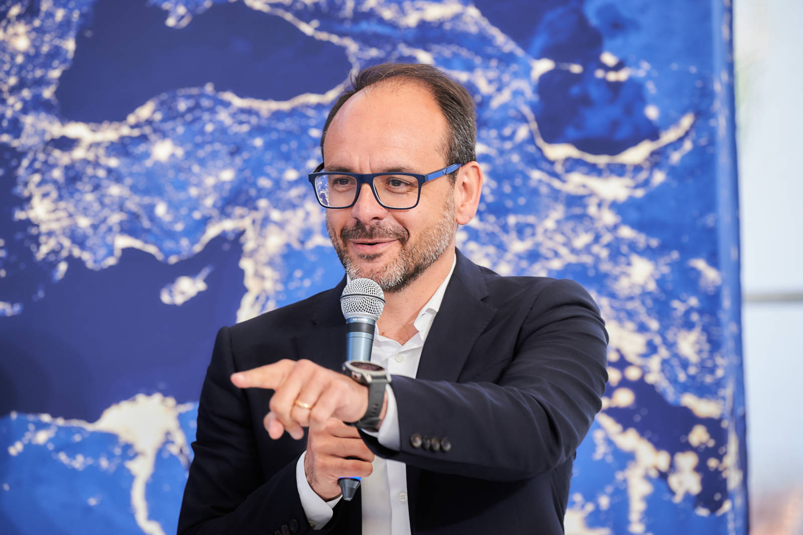 Un oratore gesticola mentre tiene un microfono su uno sfondo di una mappa del mondo astratta in blu e bianco.