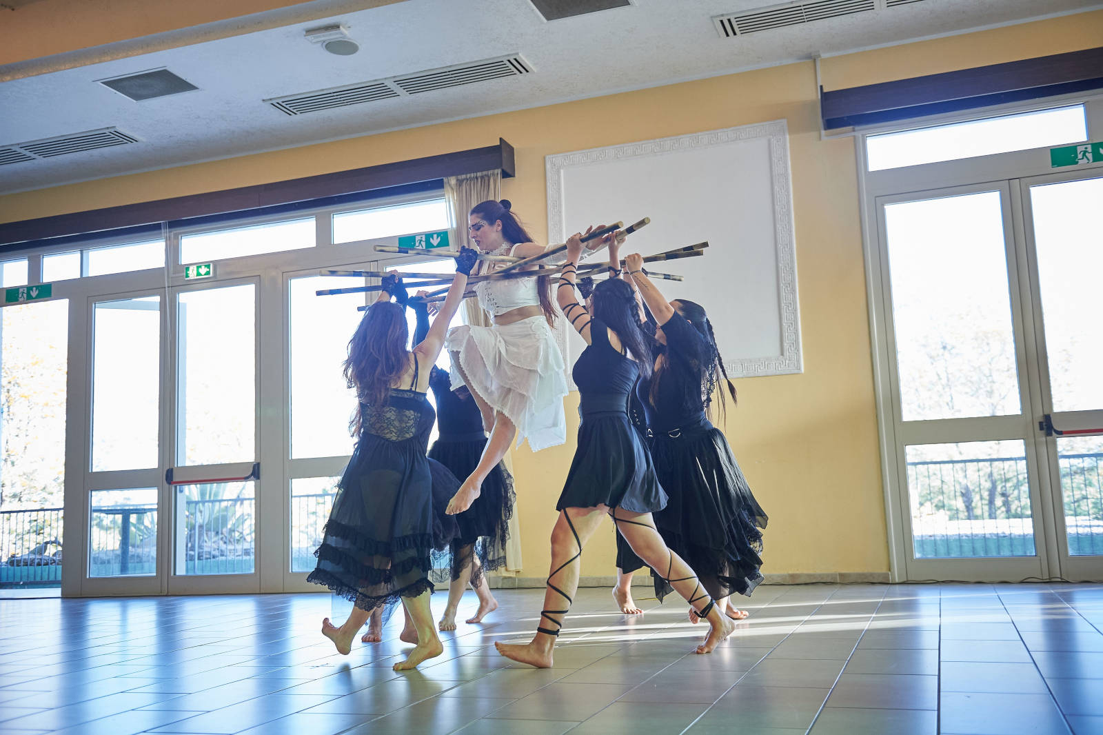 Un gruppo di ballerine in abiti neri solleva una danzatrice in un ambiente luminoso con grandi finestre.