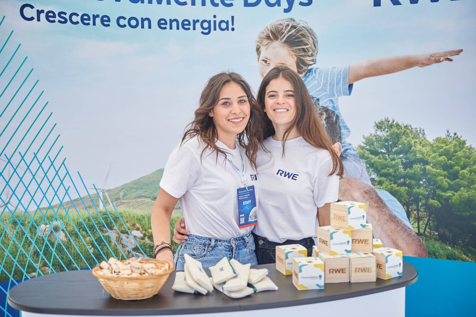Due persone in magliette bianche RWE si trovano accanto a un tavolo con articoli promozionali e un cesto di gadget.