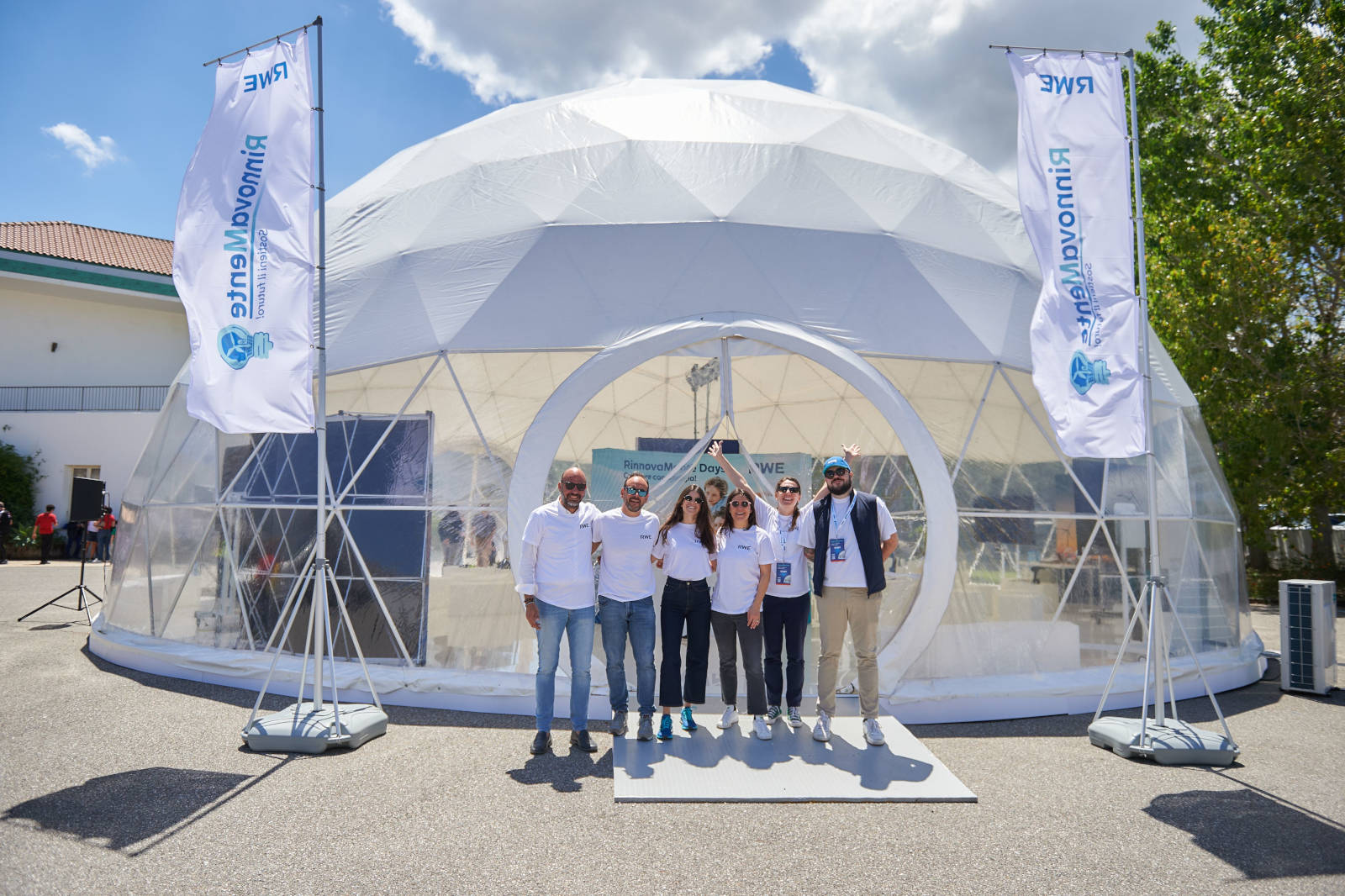 Un gruppo di persone in magliette bianche posa davanti a una cupola geodetica a un evento, con striscioni e bel tempo.