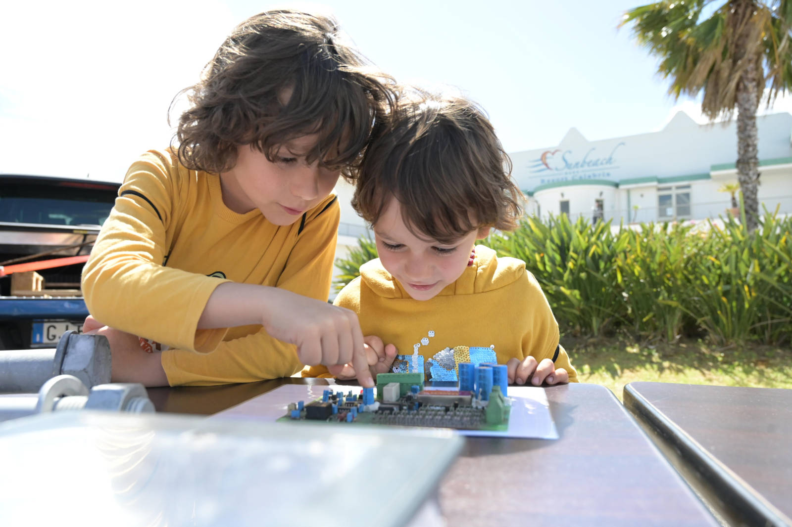 Due bambini in magliette gialle interagiscono con un piccolo modello su un tavolo, con palme e un edificio sullo sfondo.