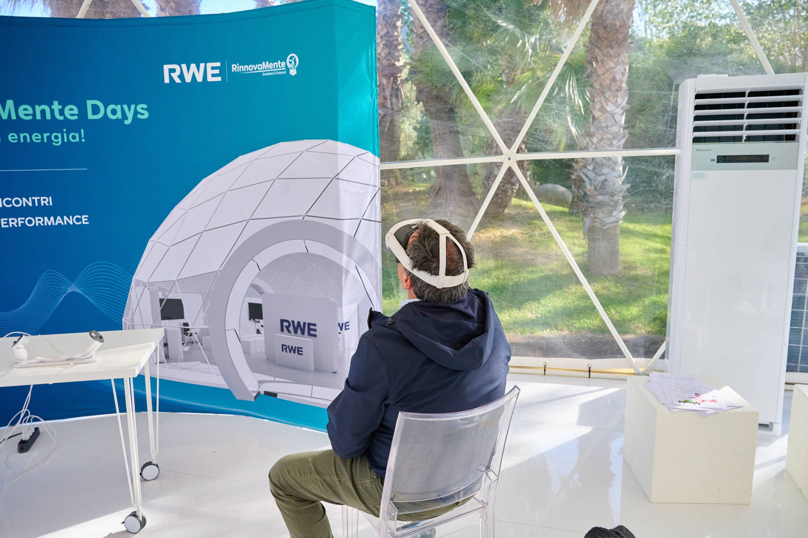 Un uomo con un visore siede davanti a uno stand promozionale di RWE, mostrando un evento energico in uno spazio luminoso e moderno.