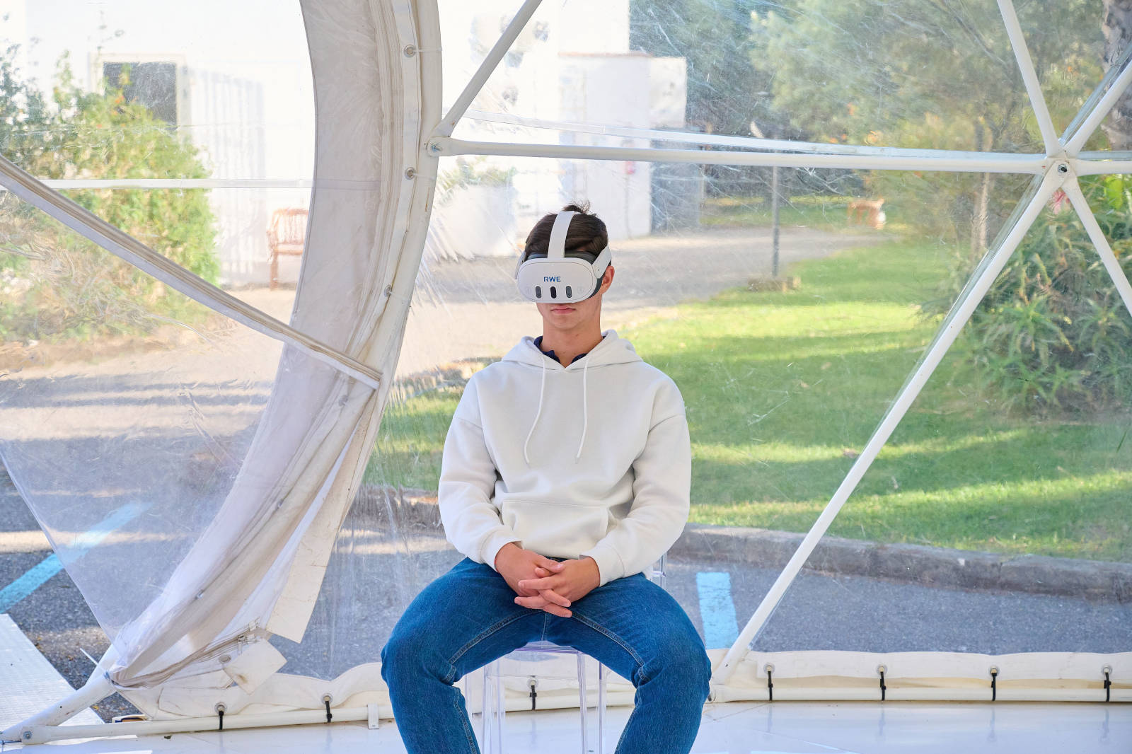 Un giovane uomo seduto dentro una struttura trasparente, indossa una felpa bianca e jeans, in un ambiente esterno.