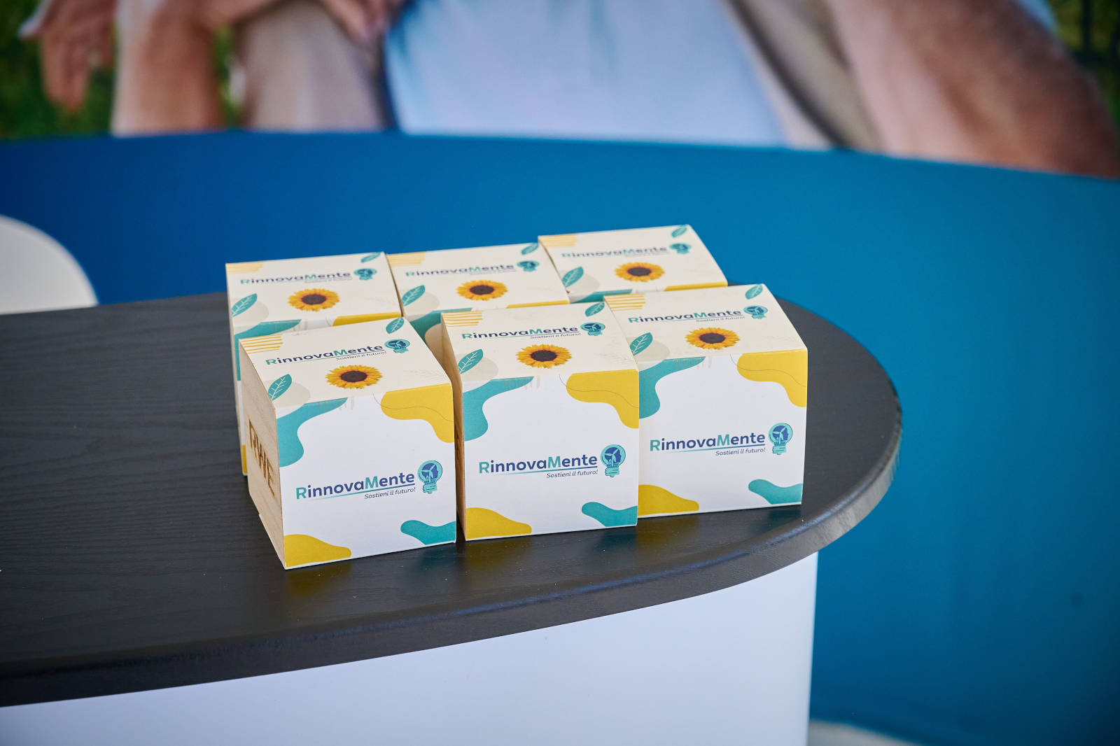 Diverse scatole di RinnovaMente esposte su un tavolo nero a un evento. Lo sfondo presenta uno striscione blu.