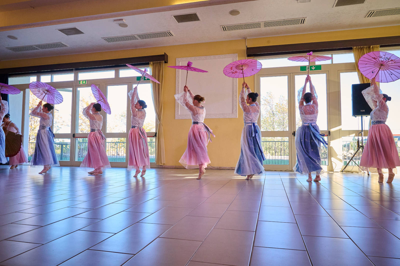 Un gruppo di ballerine in abiti colorati che si esibiscono con ombrelli rosa in un'ampia sala interna.