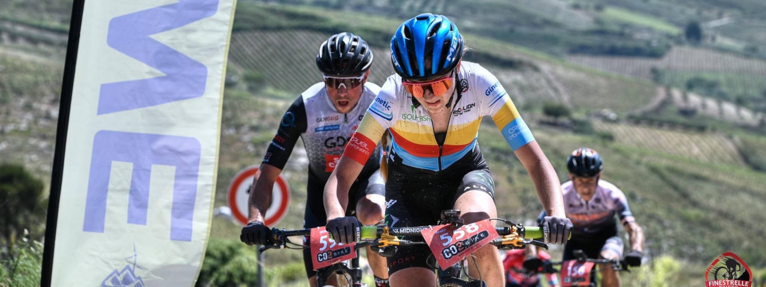 Three cyclists in a race, wearing colourful jerseys, ride through a scenic landscape with hills and vineyards.