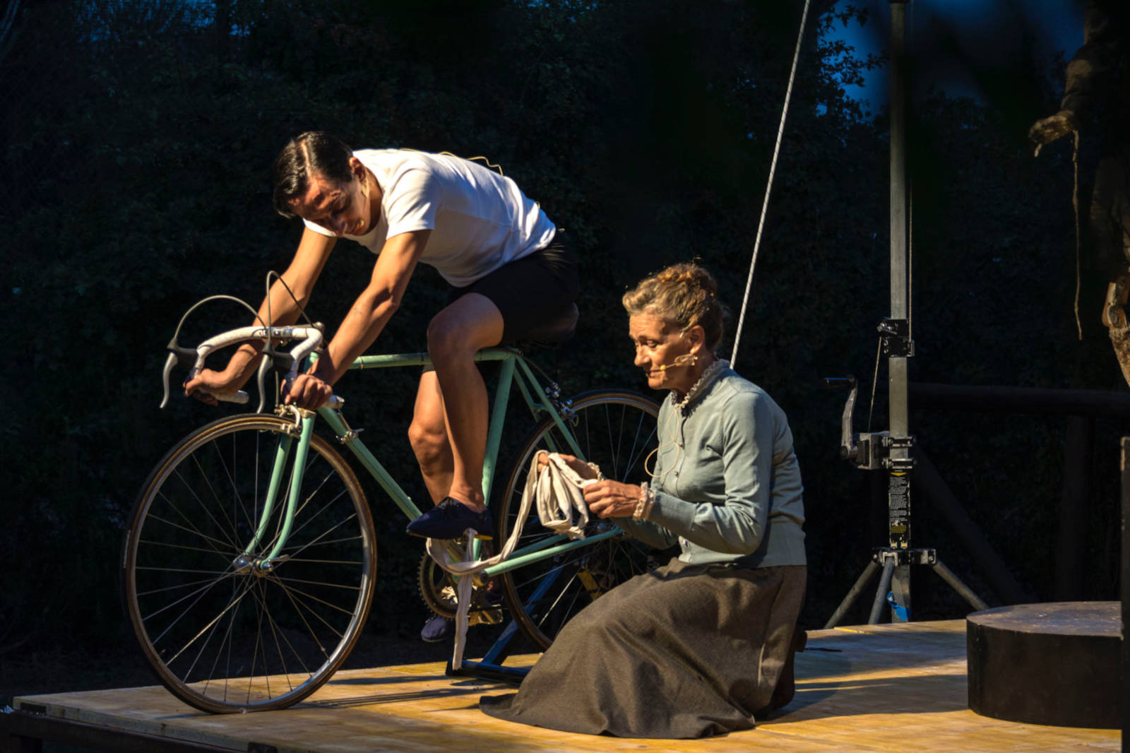 A cyclist is positioned on a stationary bike, while a seated woman assists with a cloth on a wooden stage.