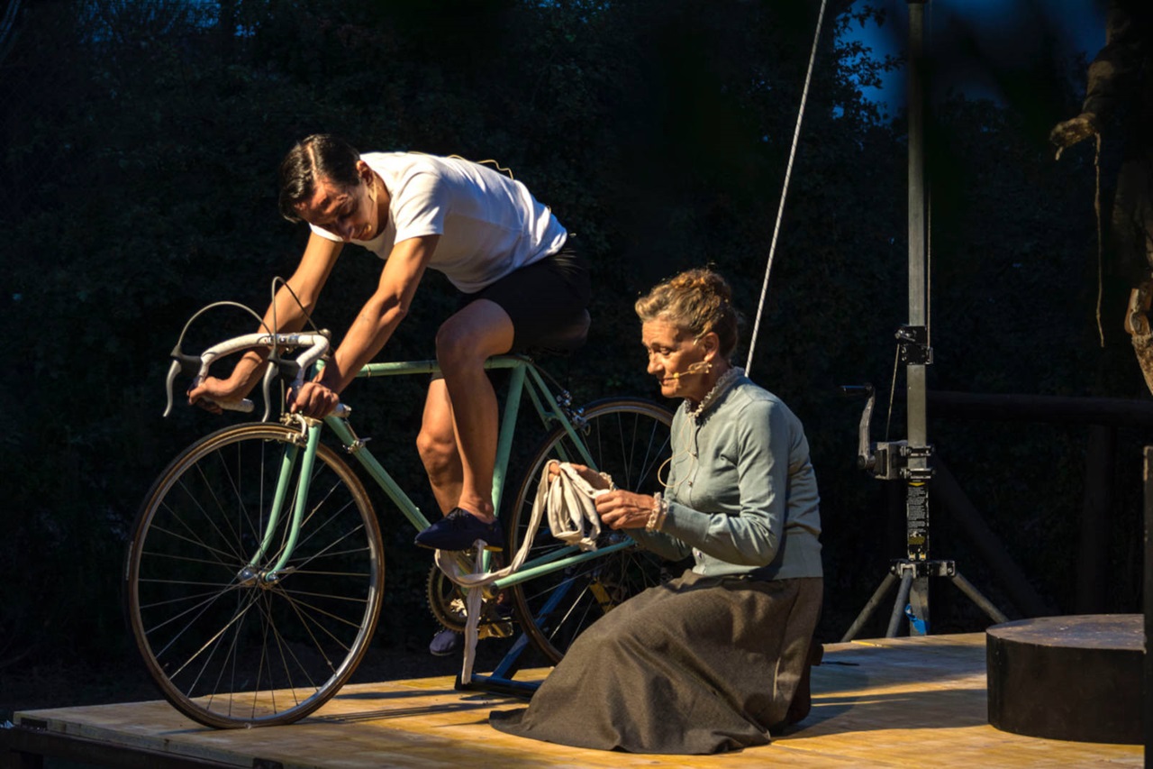 Un ciclista è posizionato su una bicicletta ferma, mentre una donna seduta aiuta con un panno su un palco in legno.