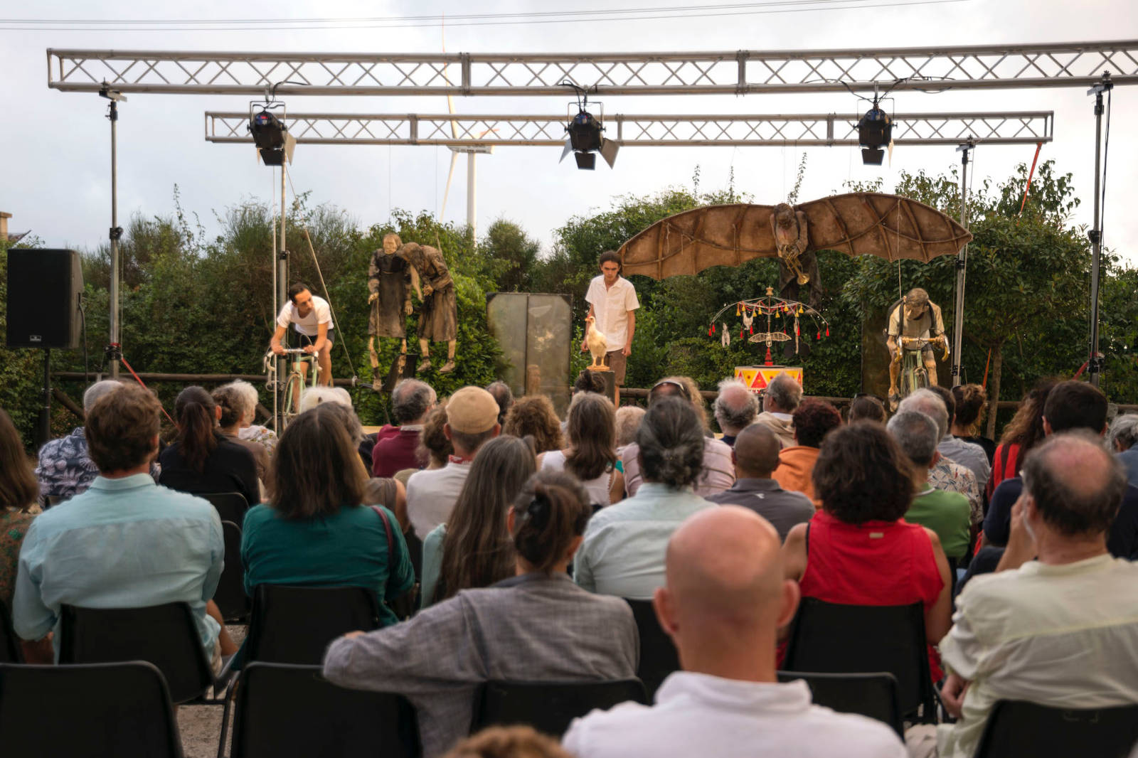 Una performance all'aperto con marionette, un pubblico eterogeneo seduto davanti, alberi sullo sfondo e luci di scena sopra.