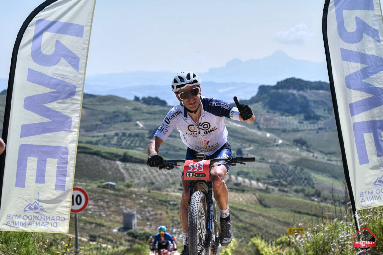A mountain biker gives a thumbs up while riding on a scenic trail, with flags and hills in the background.