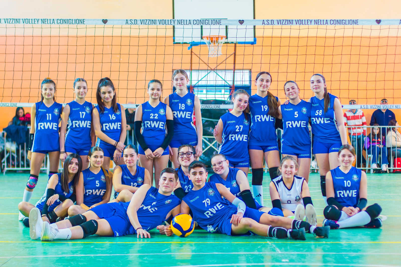 A volleyball team in blue uniforms poses on a court, with a volleyball at their feet, showcasing team spirit.