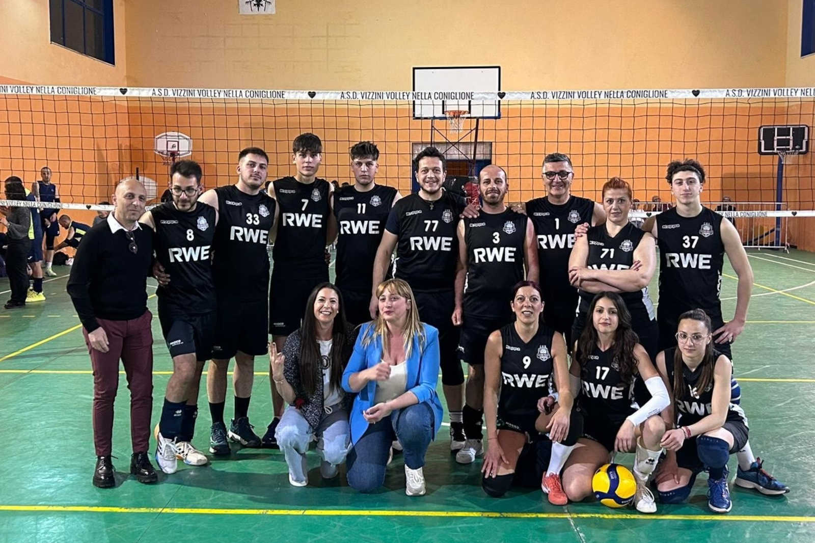 Una squadra di pallavolo posa in magliette nere con il logo RWE, con un pallone da pallavolo giallo davanti, all'interno di una palestra.