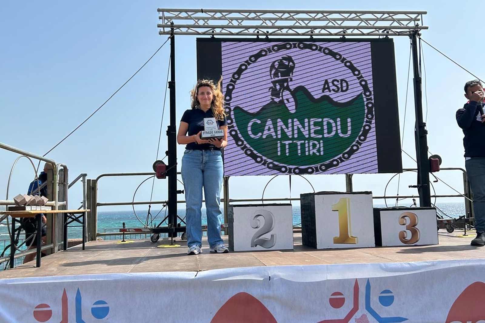 Una donna sta sul palco tenendo un premio, con uno schermo sullo sfondo che mostra il logo di Cannedu Ittiri vicino al mare.