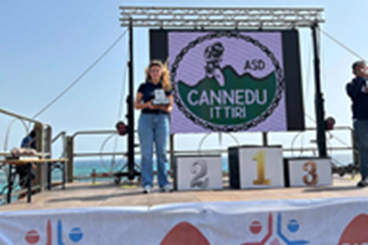 Una donna sta sul palco tenendo un premio, con uno schermo sullo sfondo che mostra il logo di Cannedu Ittiri vicino al mare.