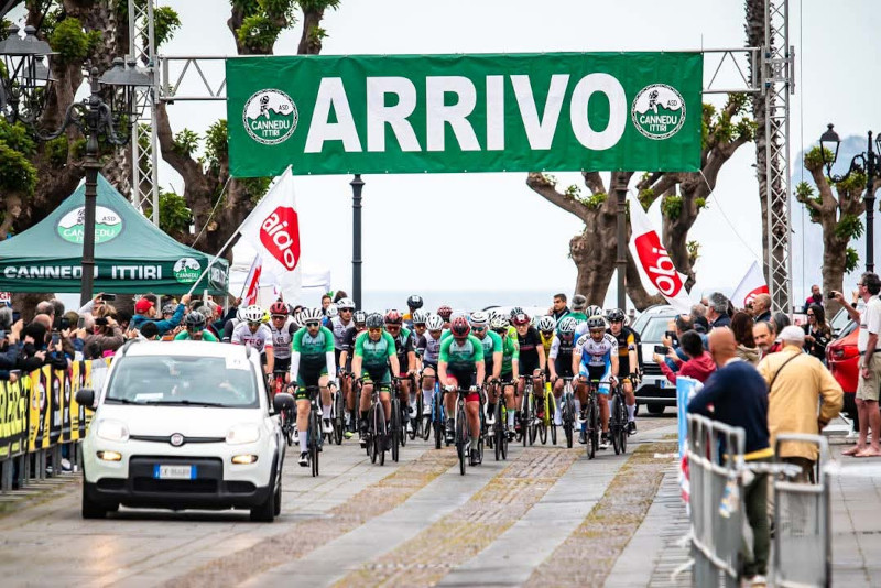 A cycling race with athletes in green and black jerseys crossing a finish line marked 'ARRIVO', surrounded by a cheering crowd.