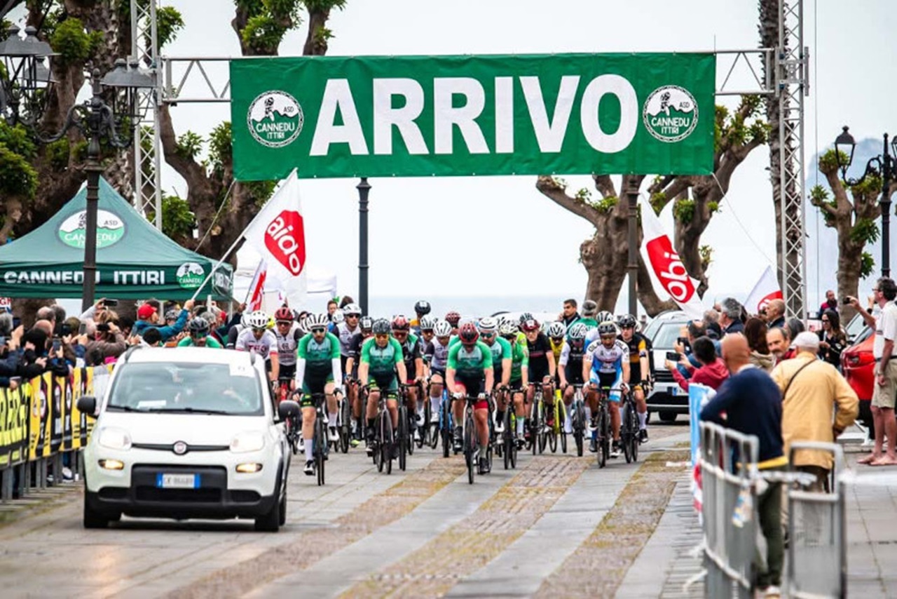 Una corsa ciclistica con atleti in maglie verdi e nere che attraversano un traguardo contrassegnato da 'ARRIVO', circondati da un pubblico festante.