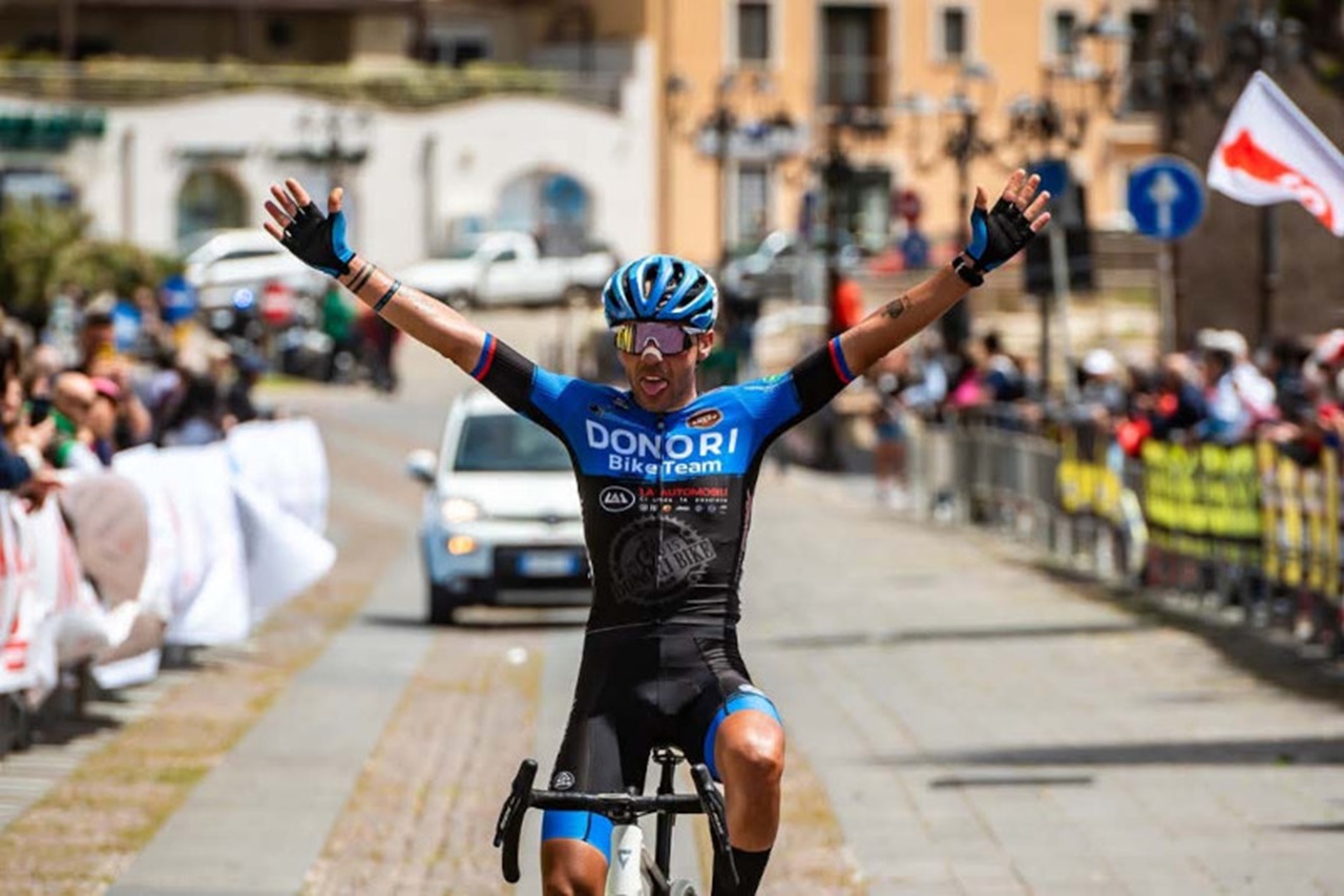 Un ciclista in una maglia blu del Donori Bike Team festeggia la vittoria con le braccia alzate su una strada piena di tifosi.