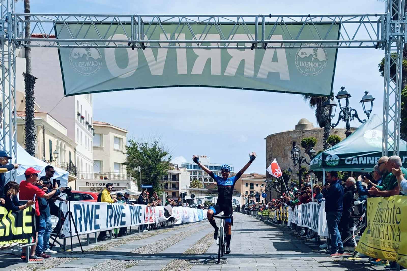 A cyclist celebrates victory at the finish line, surrounded by an enthusiastic crowd and banners.