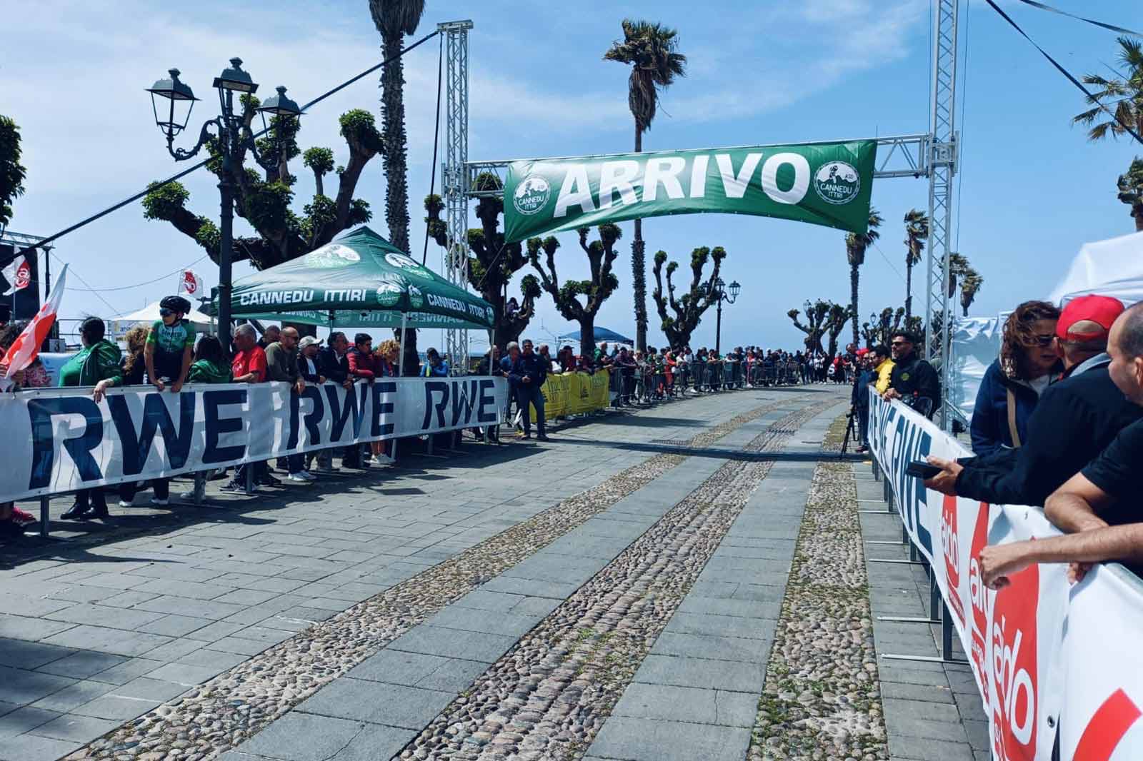 Una folla riunita al traguardo di una gara sotto un'insegna verde 'ARRIVO', con palme e tende sullo sfondo.