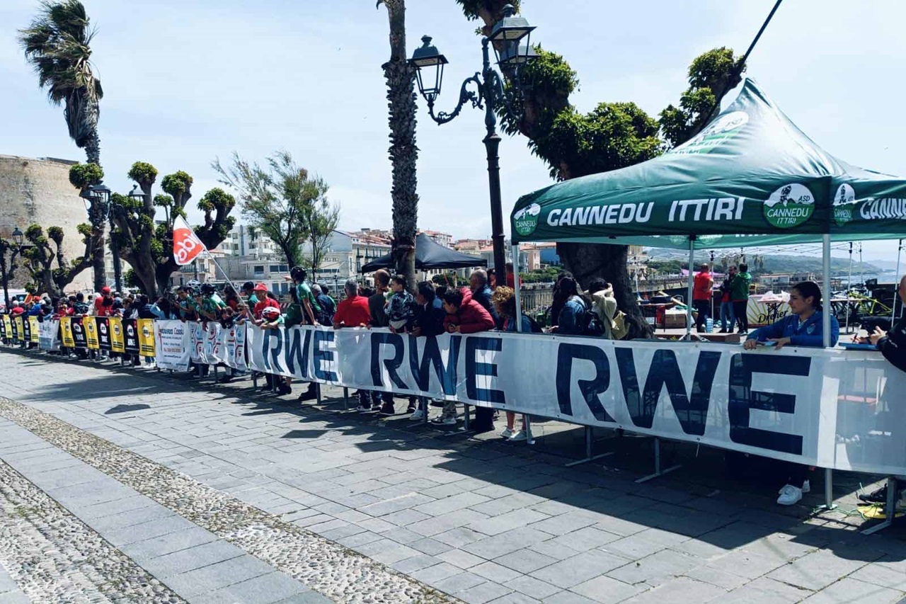 Una scena vivace di spettatori a un evento all'aperto con striscioni e una tenda che mostra 'Gannedu Ittiri' in un'atmosfera animata.