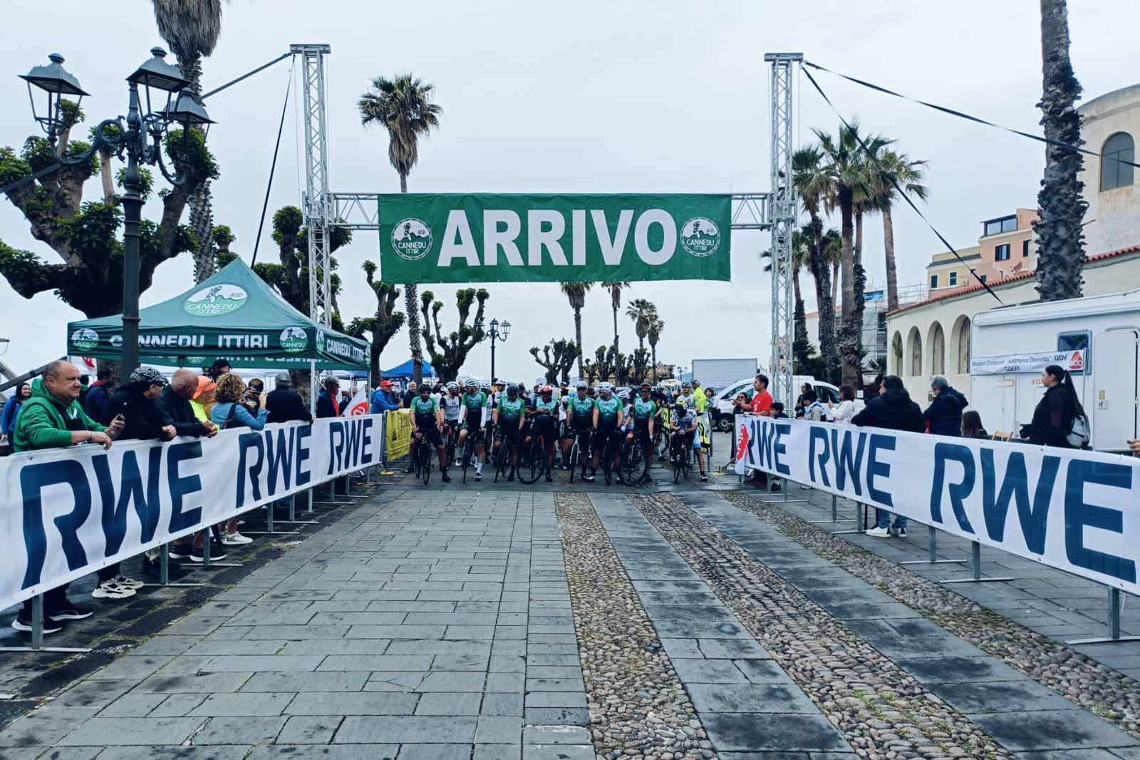 Cyclists gather at the finish line beneath a green banner labelled 'ARRIVO' at an event in a coastal setting.