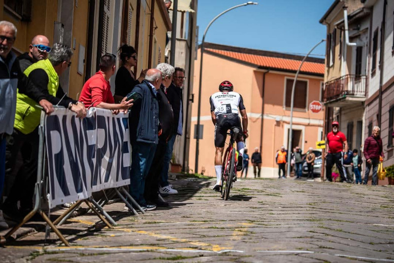 A cyclist in a black and white outfit rides past a crowd on a sunlit street, surrounded by colourful buildings.