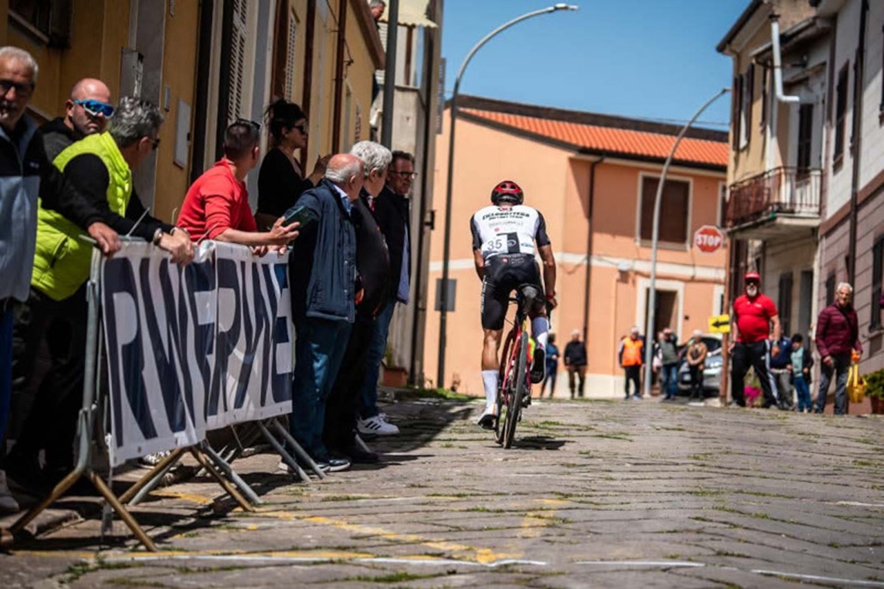 Un ciclista in un completo bianco e nero passa davanti a una folla su una strada soleggiata, circondato da edifici colorati.