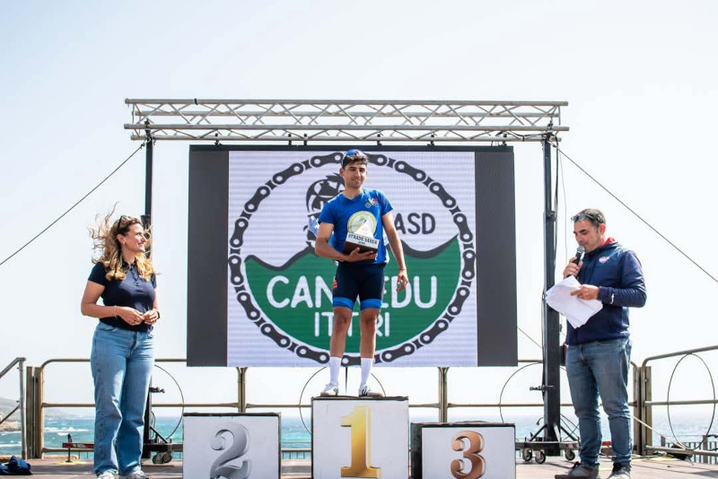 A podium ceremony with a winner holding a trophy, flanked by two presenters, set against a seaside background.