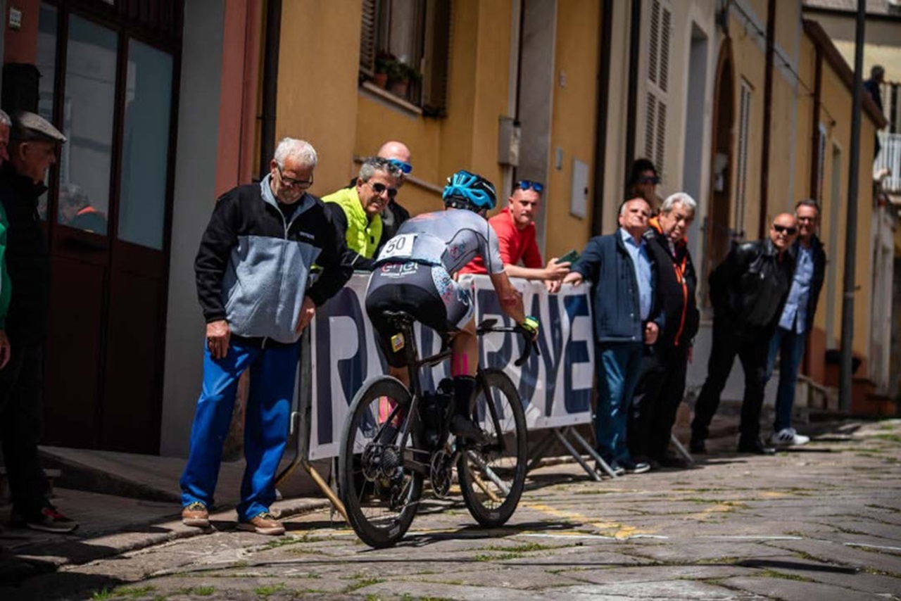 Un ciclista in abbigliamento grigio e rosa passa davanti a una folla esultante vicino a un edificio con facciate colorate.