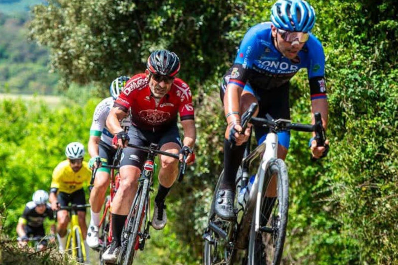Five cyclists racing on a dirt path surrounded by greenery, wearing colourful jerseys and helmets under bright sunlight.
