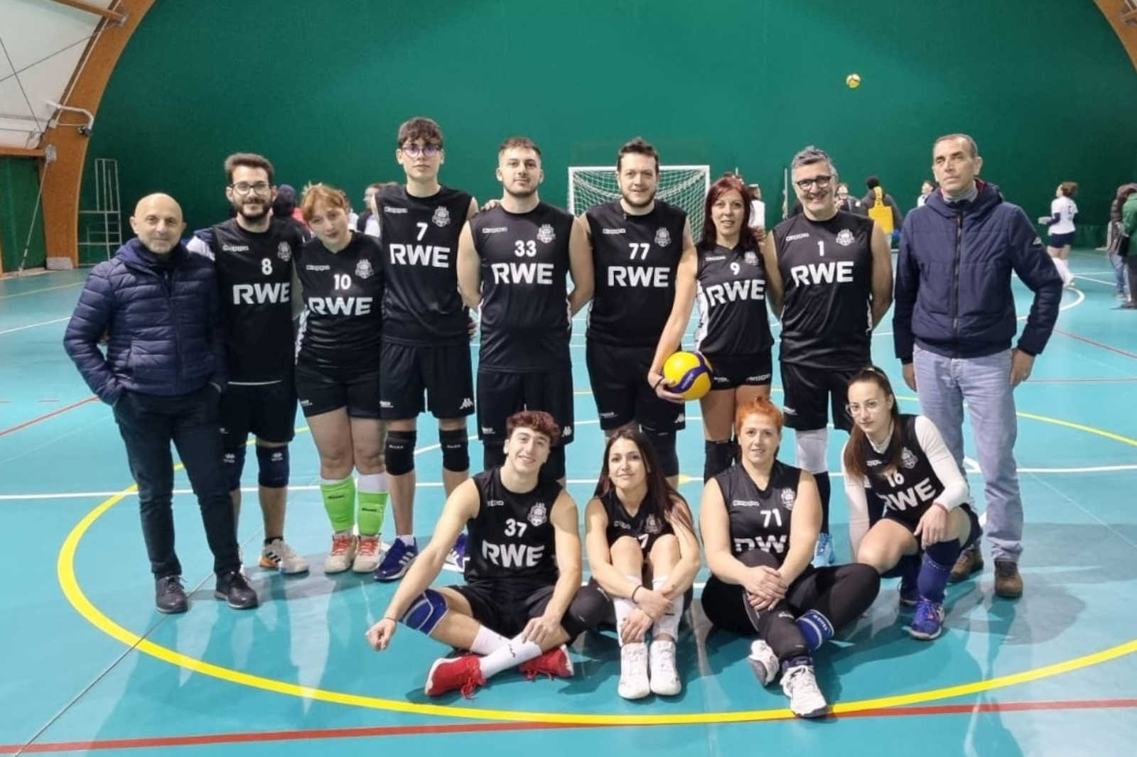 A volleyball team poses together in black jerseys with 'RWE' logo, on a gym court with a green dome ceiling.