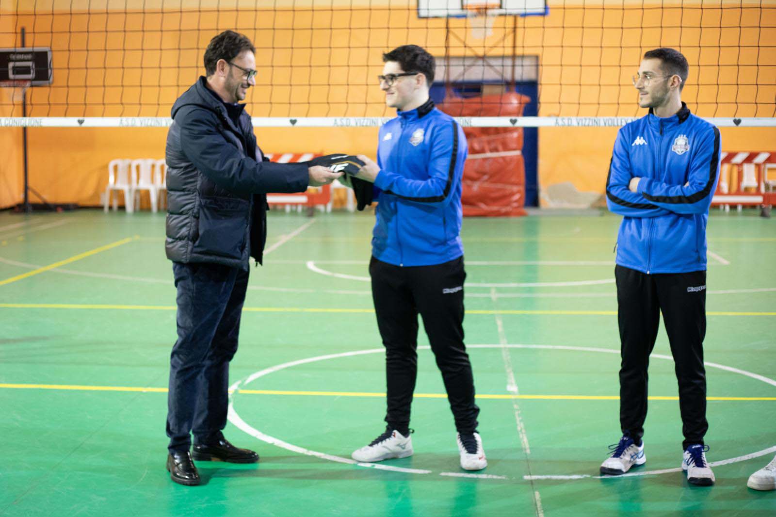 Un uomo in giacca scura consegna un premio a un atleta in tuta blu, con un altro atleta che osserva, in una palestra.