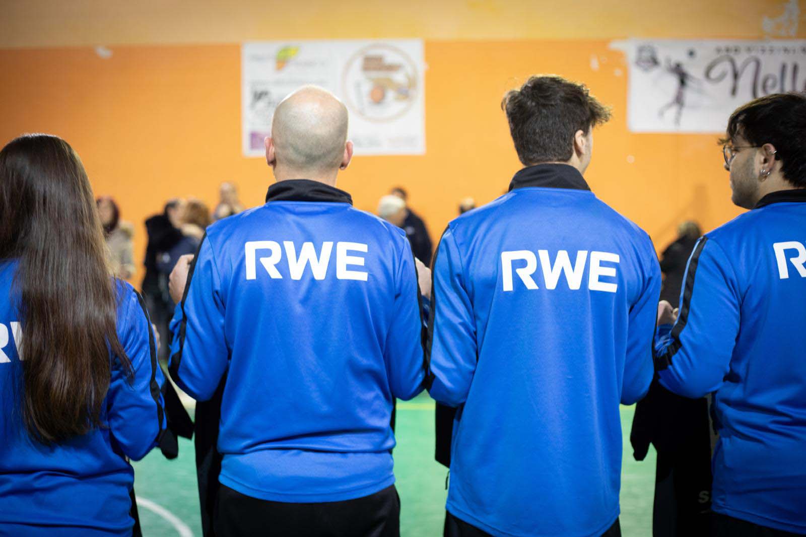 Un gruppo di individui in giacche blu coordinate, con 'RWE' sulla schiena, in piedi in una palestra.
